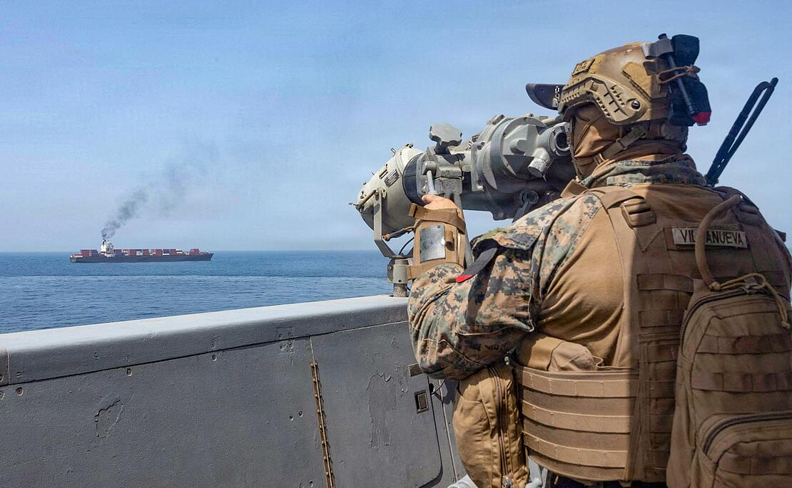 Im Vordergrund ein Soldat auf einem Schiff, der einen Öltanker im Hintergrund mit einem Fernrohr ins Visier nimmt