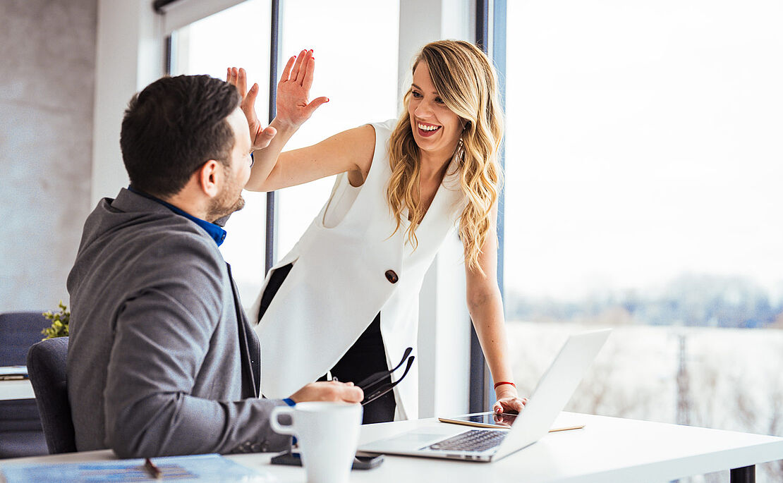 Ein Mann - sitzend am Schreibtisch - und eine Frau - stehend daneben - klatschen freudig ab