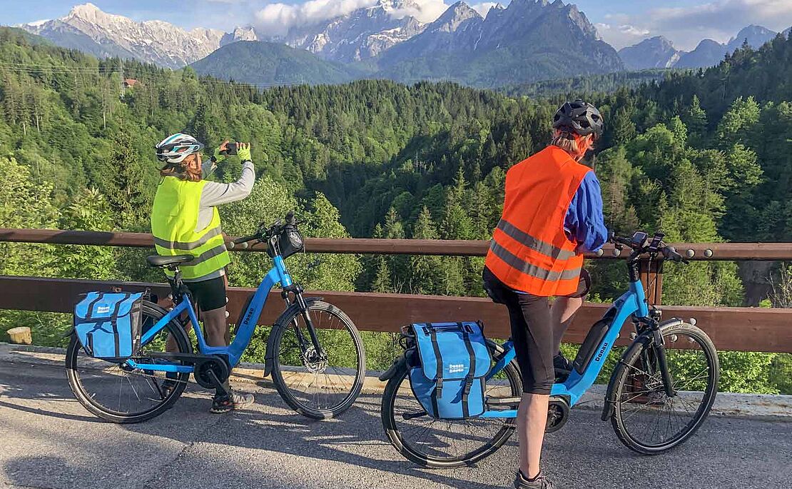 Mann und Frau auf E-Bike, Frau fotografiert die Landschaft bei Tarvis mit Blick auf die Julischen Alpen