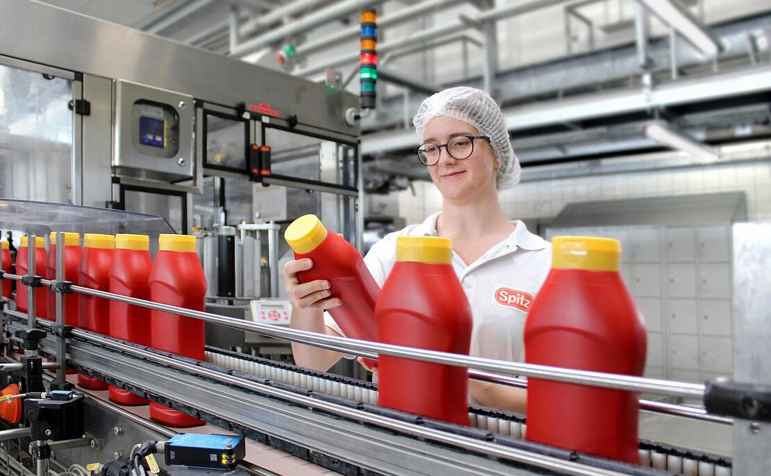 Junge Frau mit Haarnetz und weißem Shirt am Fließband mit Ketchupflaschen