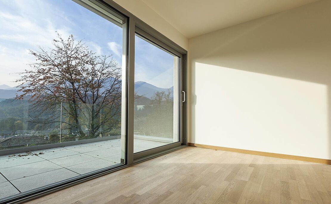 Leeres Zimmer mit Holzbelag, Blick aus den raumhohen Terrassenfenstern in einen Garten