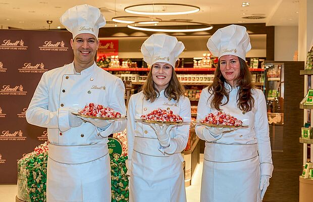 Ein Mann und zwei Frauen stehen im Kochgewand ganz in weiß und mit Kochhaube am Kopf jeder mit einem Tablett Lindt-Süßigkeiten nebeneinander