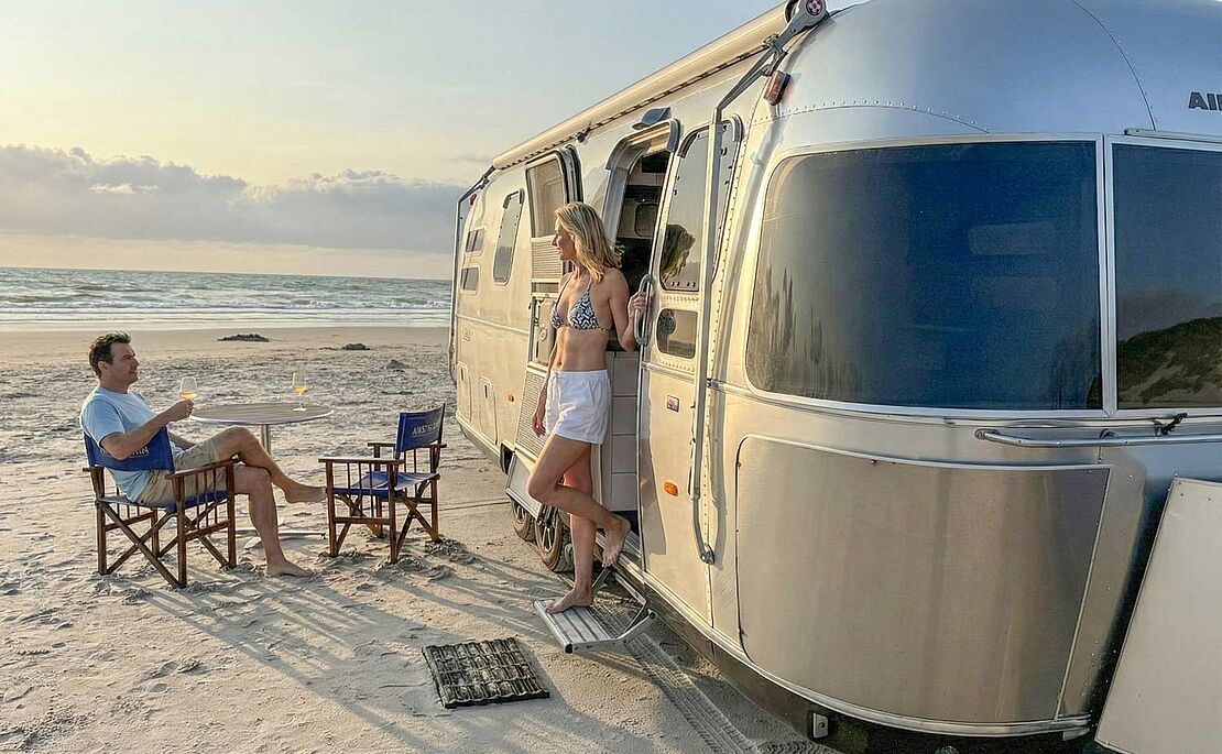 Silbernes Wohnmobil, geparkt am Strand, eine Frau im Bikini tritt gerade aus der Tür, ein Mann sitzt davor an Tisch mit zwei Sesseln
