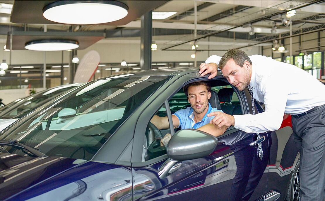 Ein Mann sitzt in einem Autohaus in einem blauen Auto, das Seitenfenster ist geöffnet, daneben stehend erklärt ihm ein anderer Mann etwas