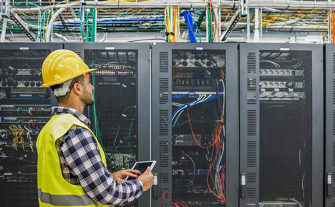 Mann in Schutzkleidung und Helm vor einem großen Schaltkasten mit Leitungen