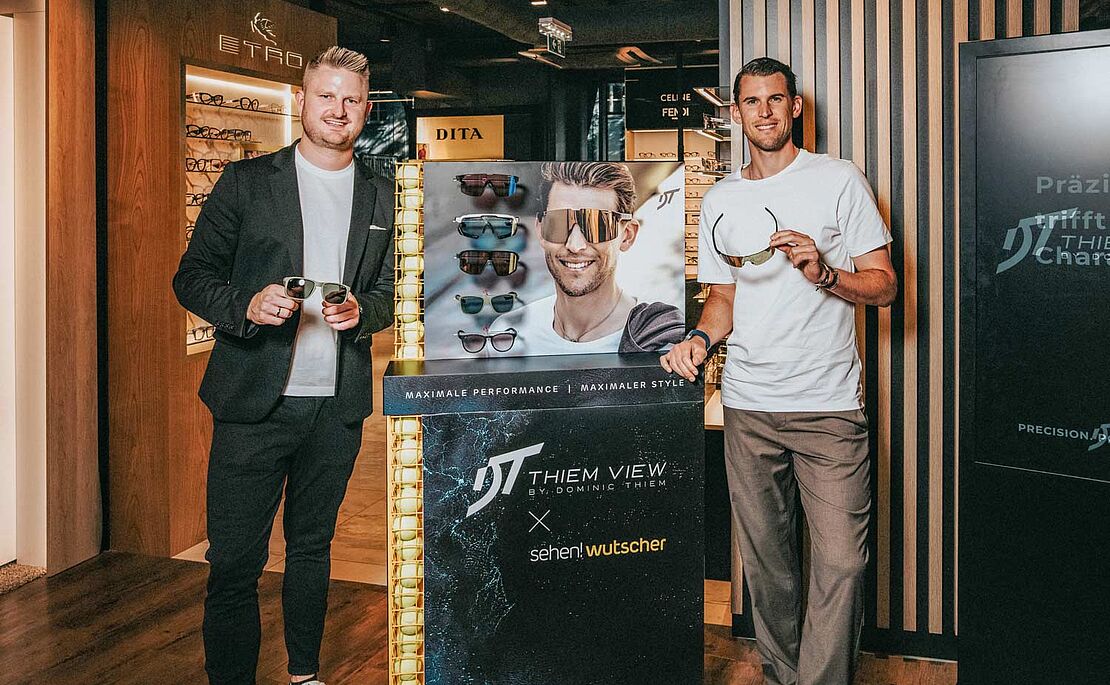 Fritz Wutscher jr. und Dominic Thiem vor einem Brillengeschäft, jeder hält eine Sonnenbrille in der Hand, in ihrer Mitte ein Werbeständer