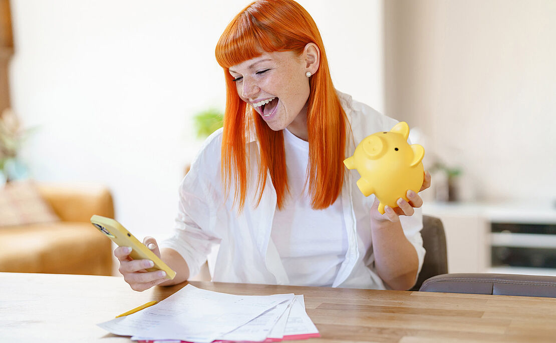 Lachende Frau mit roten Haaren und einem Handy und einem Sparschwein in der Hand