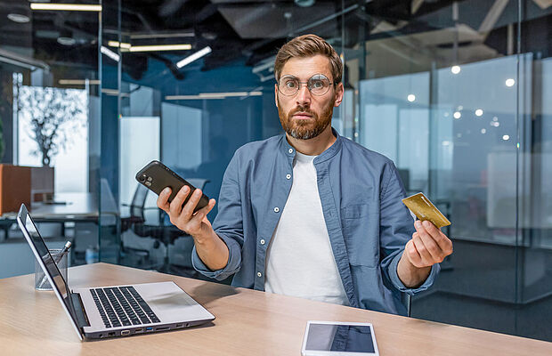Mann mit Brille und Vollbart hält in einer Hand das Handy, in der anderen eine Kreditkarte und blickt verwundert
