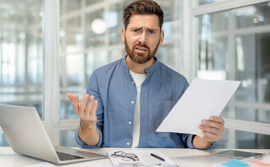 Mann vor PC und mit Papier in der Hand, der fragend und verständnislos in die Kamera schaut